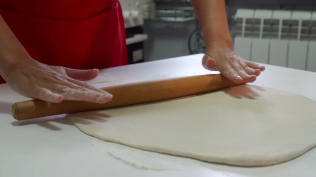 A flat sheet of white dough lies on the floured counter. A wooden rolling pin moves quickly in the cook's hands to flatten the pastry base for a delicious homemade meal.