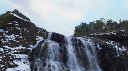 Aerial winter view of a powerful cascading waterfall flowing between snowy rocky cliffs, creating a dramatic cinematic landscape with cold mist, frozen textures, and natural wilderness. - Powered by Adobe