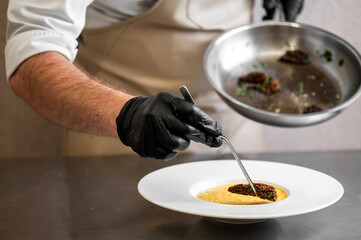 A chef wearing black gloves carefully plates a gourmet dish of yellow puree and a seared item in a white bowl, preparing food in a professional kitchen.