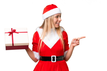 Young blonde woman with christmas hat and holding a gift over isolated chroma key background pointing to the side to present a product