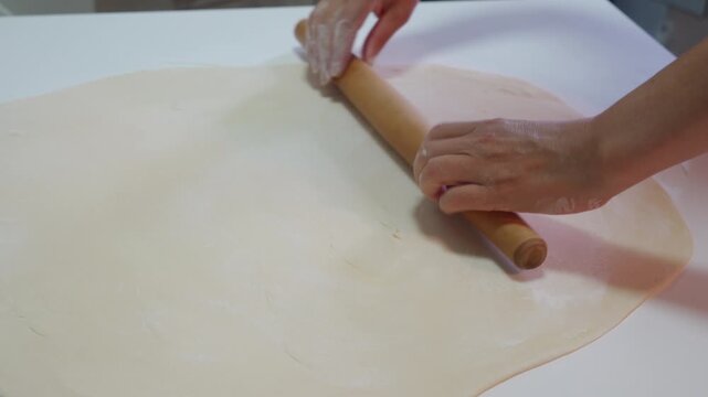 Soft floury dough is smoothed by a wooden rolling pin on a white surface. Skilled hands guide the tool to flatten the raw crust for baking a delicious meal.