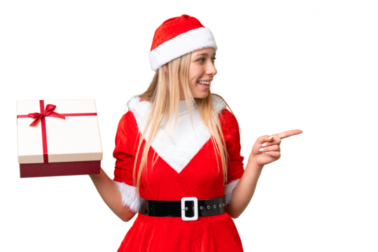 Young blonde woman with christmas hat and holding a gift over isolated chroma key background pointing to the side to present a product - Powered by Adobe
