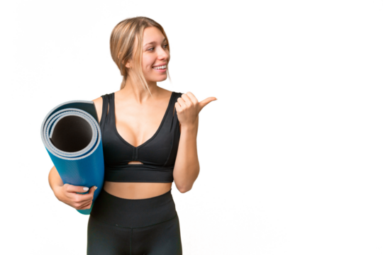 Blonde sport woman going to yoga classes while holding a mat over isolated chroma key background pointing to the side to present a product