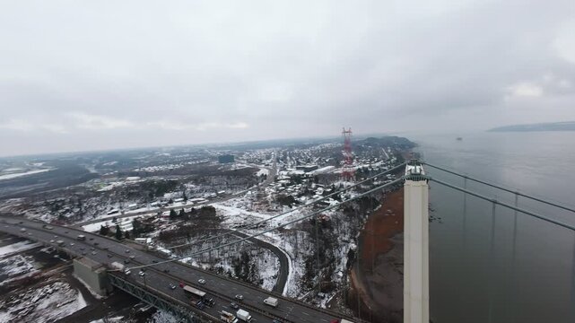 Aerial cinematic drone view of a large steel bridge crossing a wide river in winter. Snowy shoreline, cloudy sky and industrial architecture create a dramatic cold season atmosphere.