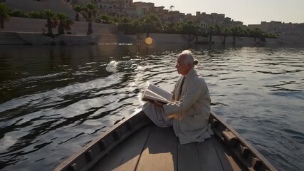 Man reads book on boat. Senior sits quietly near river at sunset. Elderly man reflects while reading. Calm water ripples around boat. Travel and solitude captured in natural light. Peaceful moment.