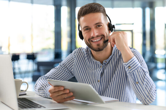 Cheerful young support phone male operator in headset, at workplace while using digital tablet