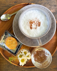 Overhead view of a frothy cappuccino served with a glass of water, delicate white daisy flowers, and a cracker arranged neatly on a warm wooden tray. Soft blurred background creates a calm, cozy café 
