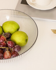 Minimalist still life of green pears and red grapes in ribbed glass bowl on white table with magazine.
