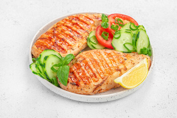 Grilled chicken breast served with fresh cucumber, tomato slices, basil and lemon on a white plate top view