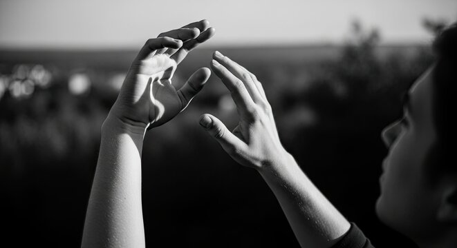 A black and white artistic photo of a person's expressive hands raised in a gentle gesture against a blurred natural background, evoking a contemplative and spiritual mood