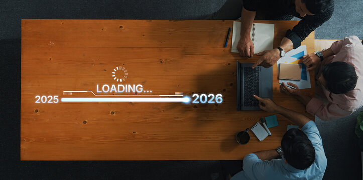 A diverse group of professionals collaborates at a wooden table, focusing on a digital project visualization showing a loading progress bar between 2025 and 2026. Trope