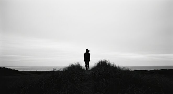 A dramatic black and white panoramic view of a lone woman's silhouette standing on a grassy sand dune, looking out at the ocean on a windy, overcast day