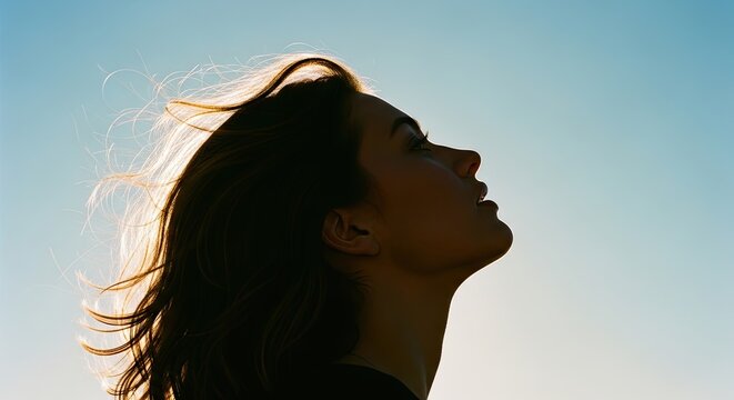 A serene woman's face in profile looking up towards the light with a hopeful expression, her hair blowing in the wind and backlit by the beautiful morning sun