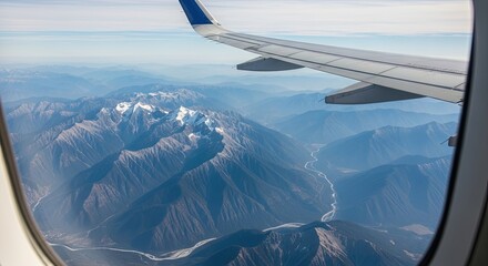 Obraz premium Breathtaking aerial view from an airplane window over a majestic mountain range with snow-capped peaks and a winding river, seen past the aircraft wing on a clear sunny day