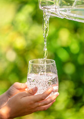 Fresh drinking water pouring into glass outdoors with human hands holding the cup