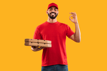 A cheerful delivery worker in a red shirt and cap stands against a vibrant yellow background. He...