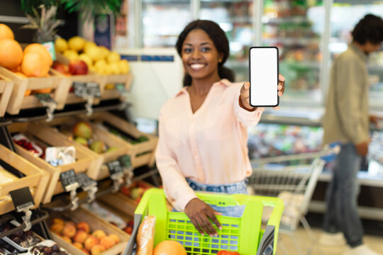 A woman smiles while showing her phone, likely with a shopping list, in a grocery store. Shelves filled with fresh fruits and vegetables are visible around her.