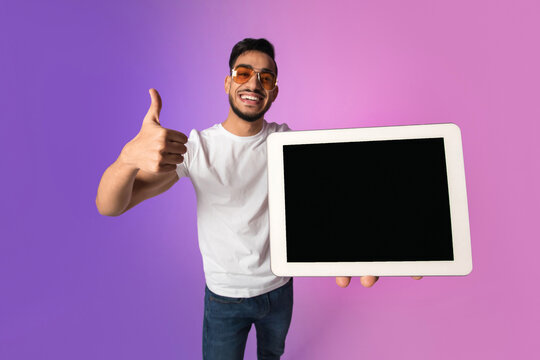 A cheerful young Arab man is presenting a tablet with an empty screen, giving a thumbs up while suggesting a new application or website. The colorful neon background enhances the vibe.