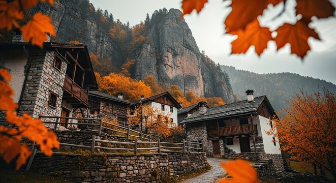 Rustic stone and wood houses of a tranquil mountain village surrounded by majestic cliffs and colorful autumn trees on a moody, overcast day, framed by vibrant orange leaves - Powered by Adobe