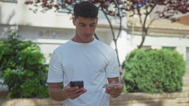 Young hispanic man holding smartphone and creditcard while looking at screen in green park outdoors; mobile payment convenience.