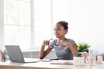 A happy young black girl smiles as she takes off her glasses, enjoying a moment of rest from studying at her laptop in her bright living room. She reflects on her upcoming exam.