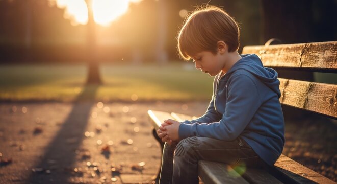 Little boy child sitting alone on a park bench looking down in sadness, demonstrating feelings of autism spectrum disorder, developmental delay