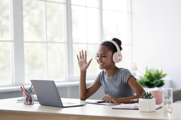 An afro American girl smiles and waves during a video call on her laptop. She is studying at home, engaged in remote learning with bright natural light coming through the windows.