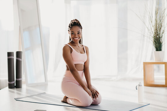 A smiling young black woman takes a break from her home workout, resting on a yoga mat. She practices fitness indoors during the covid lockdown, showcasing a positive attitude.