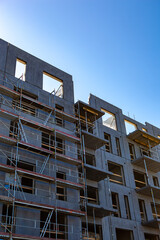Workers are installing scaffolding and completing exterior walls of a new building. The construction site is bustling with activity under a bright blue sky, indicating progress