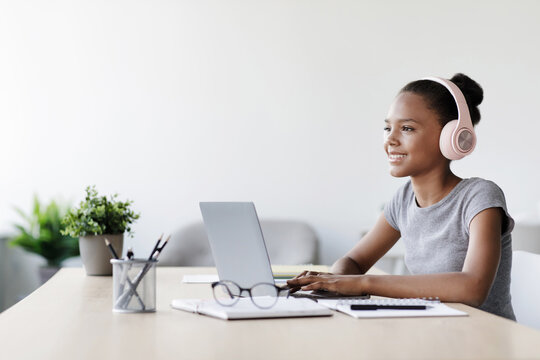 Happy teen girl listens to video lesson through headphones while focusing on her laptop in a bright living room. This scene reflects modern education during the COVID-19 quarantine.