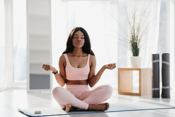 A beautiful young black woman sits in lotus pose on her yoga mat at home, eyes closed, immersed in...