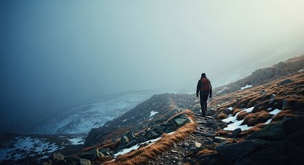 A lone hiker with a backpack walks along a rocky mountain trail through a vast, snowy landscape on a cold and foggy day, embodying adventure and solitude