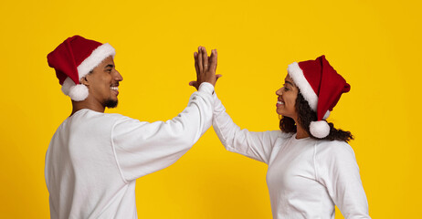 A man and woman are smiling and giving each other a high five while wearing red Christmas hats. The bright yellow background adds a festive vibe to their joyful interaction.