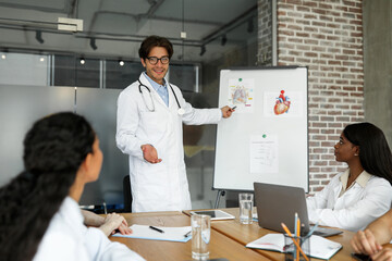 Ceerful young man chief physician standing by presentation board, having speech, multiracial team of professional doctors attending medical conference, listening to speaker, using gadgets