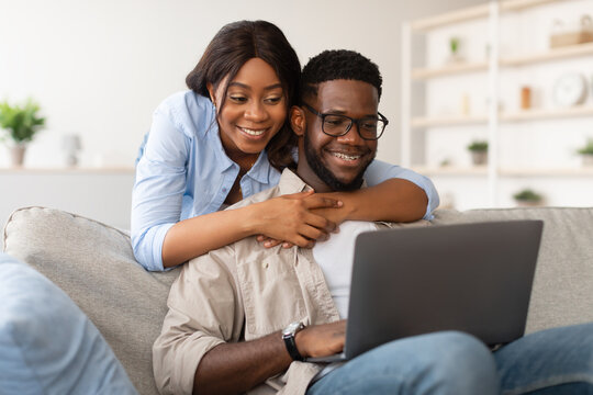 Young black couple smiles as the lady embraces her partner from behind while he works on his laptop. They are sitting on a cozy sofa in a warm home environment.