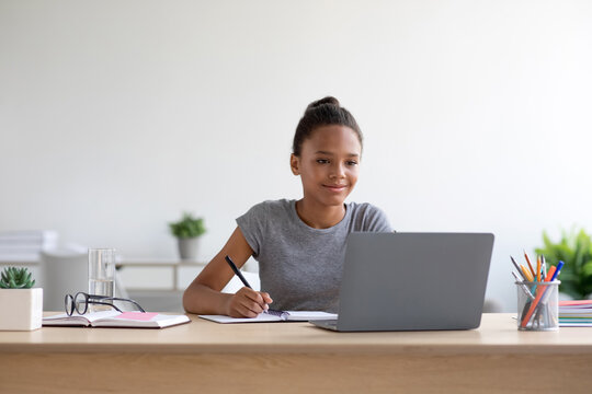 Smart young african american girl studies at home in a cozy living room, wearing headphones and using a laptop. She is focused on her homework while preparing for upcoming tests.