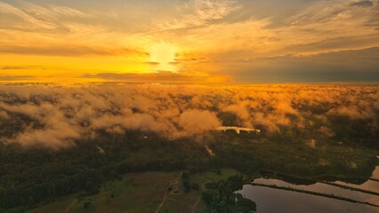 A high-altitude aerial photograph capturing a sunrise above a vast forest with golden morning mist drifting through the treetops