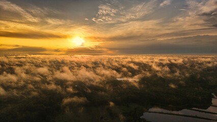 A high-altitude aerial photograph capturing a sunrise above a vast forest with golden morning mist drifting through the treetops