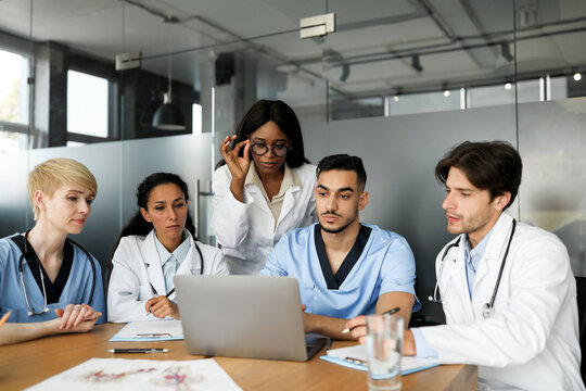 Professional multiracial team of doctors concentrated men and women in workwerar sitting at table, looking at notebook screen, taking notes, looking for solutions, clinic interior
