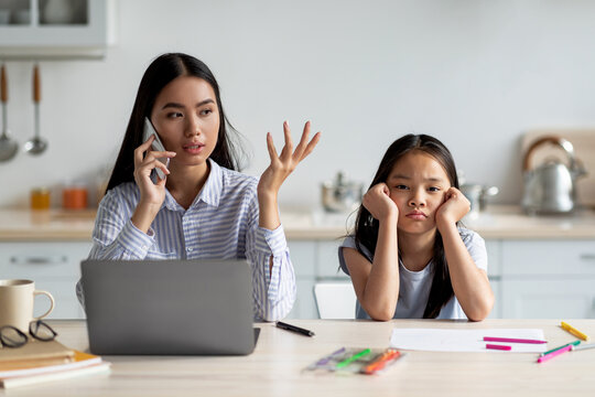 A mother sits in the kitchen answering a phone call while her daughter looks on, feeling neglected. The busy working environment highlights the challenges of parenting during lockdown.