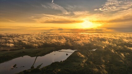 A high-altitude aerial photograph capturing a sunrise above a vast forest with golden morning mist drifting through the treetops