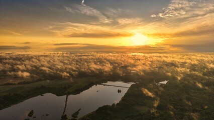 A high-altitude aerial photograph capturing a sunrise above a vast forest with golden morning mist drifting through the treetops