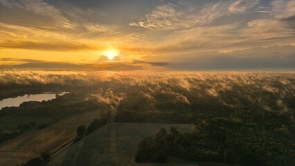 A high-altitude aerial photograph capturing a sunrise above a vast forest with golden morning mist drifting through the treetops