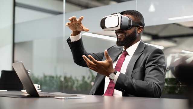 Bearded young black businessman testing new technologies for business, sitting at table in front of modern laptop, using VR smartglasses, gesturing and smiling, office interior, panorama, copy space - Powered by Adobe