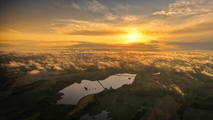 A high-altitude aerial photograph capturing a sunrise above a vast forest with golden morning mist drifting through the treetops