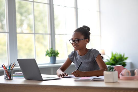 Smiling black girl in glasses studies at home with her laptop. She takes notes while learning from an online lesson, surrounded by plants and a bright window. It highlights modern education.