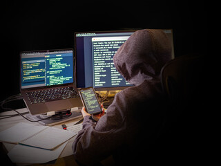 A person in a hoodie sits in front of two monitors and a laptop, displaying lines of code and a terminal. He holds a smartphone, symbolizing programming, cybersecurity, and remote work in the IT