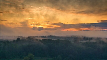 A high-altitude aerial photograph capturing a sunrise above a vast forest with golden morning mist drifting through the treetops