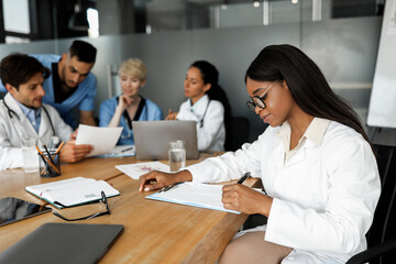 African american woman physician attending morning breefing at clinic, having conversation with her international colleagues looking at laptop screen and reports, discussing cases, taking notes