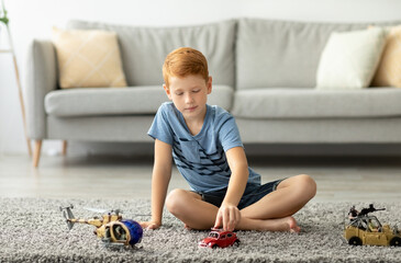 A cheerful redhead boy sits on the carpet, playing with a vintage red car and military helicopter....
